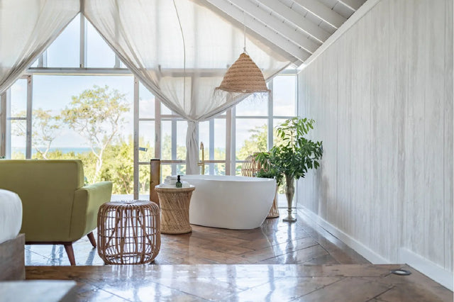 A bright, modern bathroom with a white freestanding bathtub, a green armchair, woven side tables, and large windows draped with sheer white curtains, overlooking a garden view.