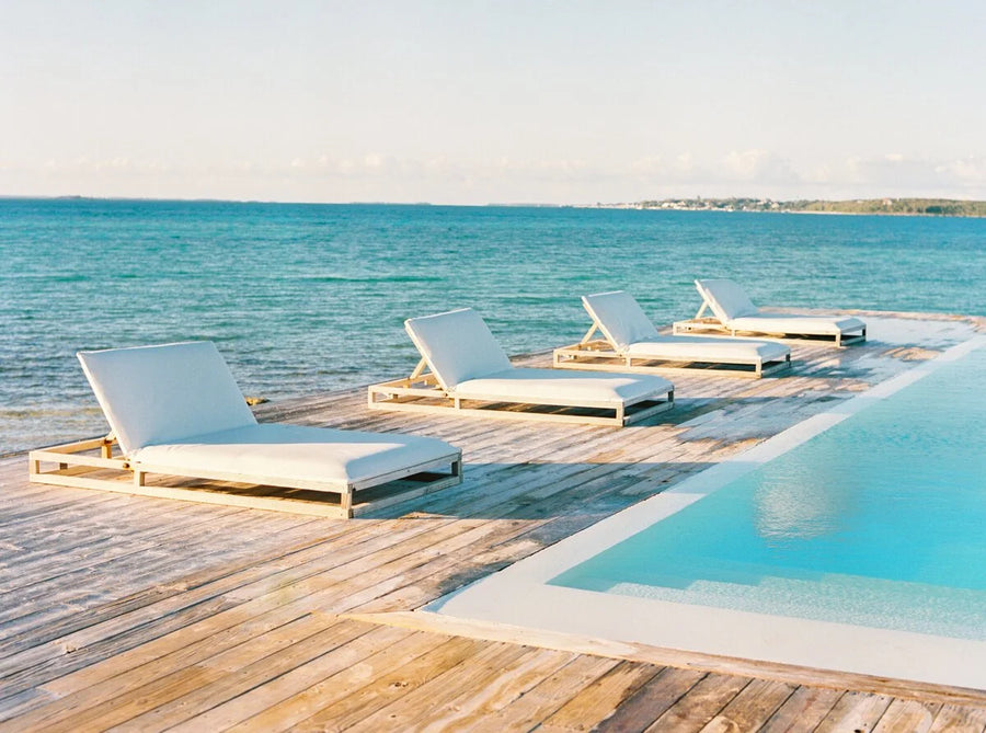 Four white cushioned lounge chairs arranged on a wooden deck beside a swimming pool with a turquoise ocean view in the background.