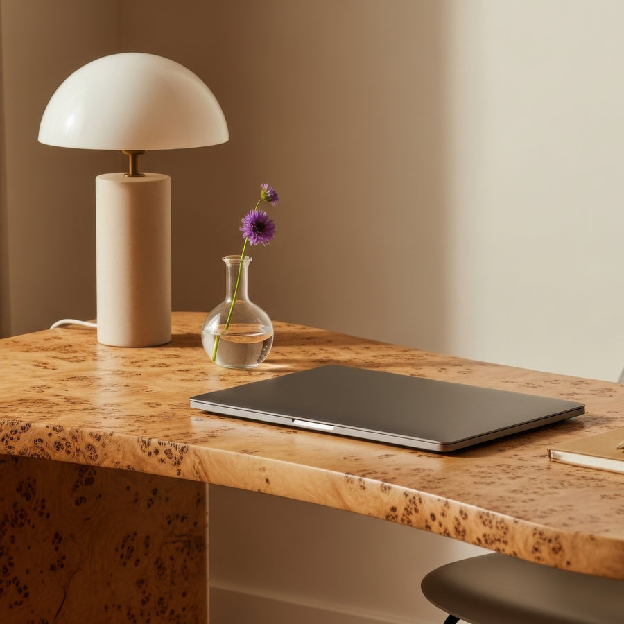 A modern desk setup featuring a beige table lamp with a white dome shade, a small glass vase with a purple flower, and a closed dark gray laptop on a burl wood desk.