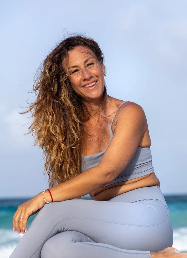 A woman with long, wavy brown hair sits on a beach wearing a matching light gray athletic crop top and leggings set.