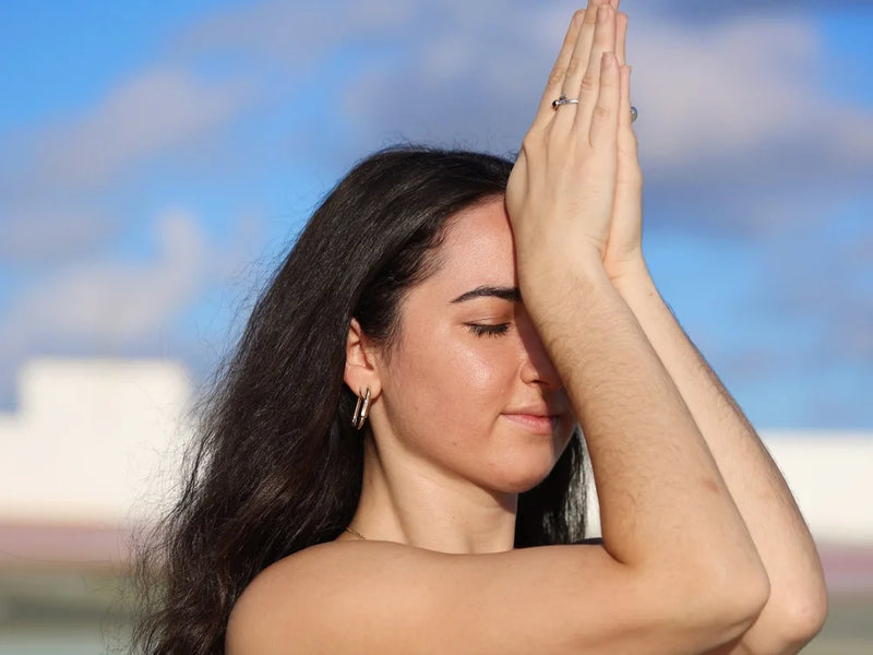 A woman with long dark hair is shown in profile outdoors with her hands pressed together over her face, wearing small hoop earrings and rings, against a blurred blue sky background.