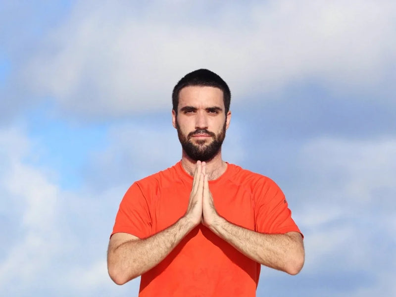 A man with a beard and short dark hair wearing a bright orange t-shirt, standing outdoors with his hands pressed together in a prayer pose against a cloudy blue sky.
