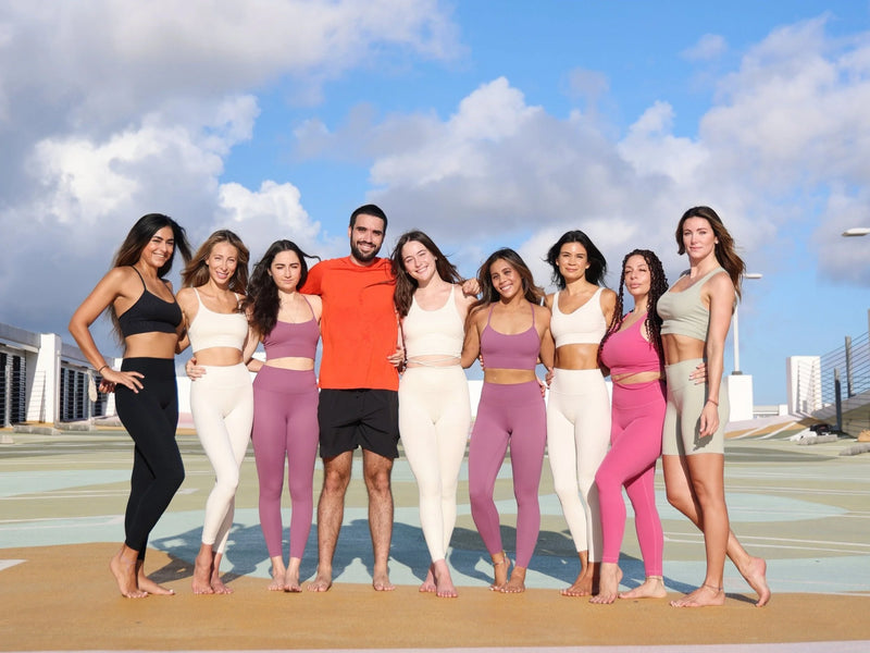 A group of nine people posing together outdoors on a sunny day, wearing matching athletic wear in various colors including black, white, purple, pink, orange, and beige. The background shows a blue sky with clouds and a modern outdoor court or plaza.