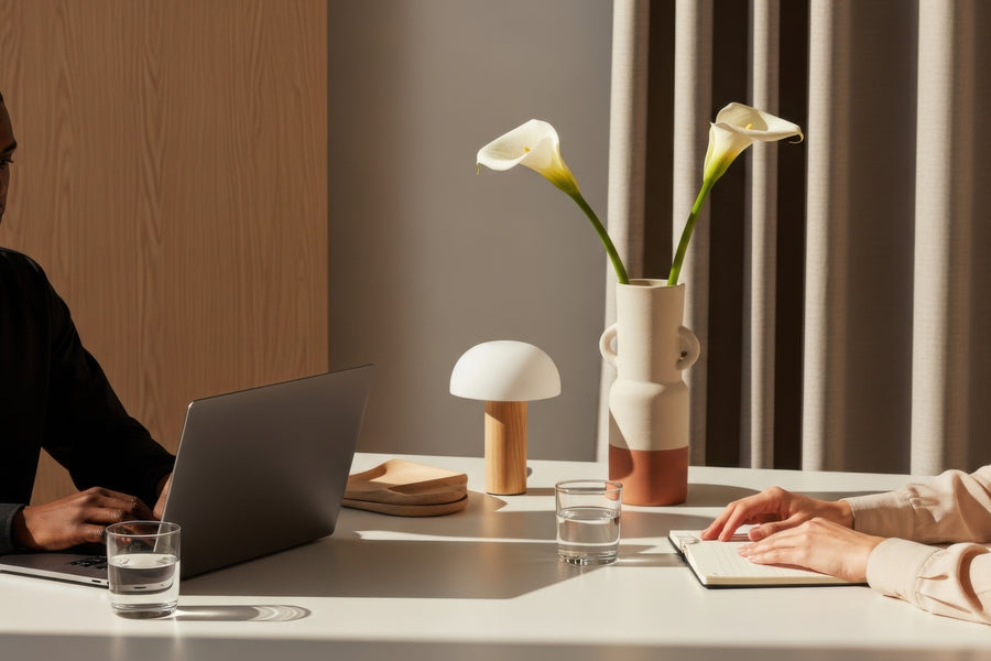 A modern interior scene with a white table holding a laptop, glasses of water, a small mushroom-shaped lamp, a ceramic vase with two white calla lilies, and a notebook. Parts of two people are visible at the table, and the background includes wood paneling and curtains.