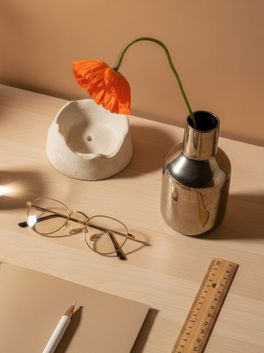 A styled tabletop scene with a metallic silver vase holding an orange flower, a white ceramic bowl, eyeglasses, a notebook, a pen, and a wooden ruler against a warm beige background.