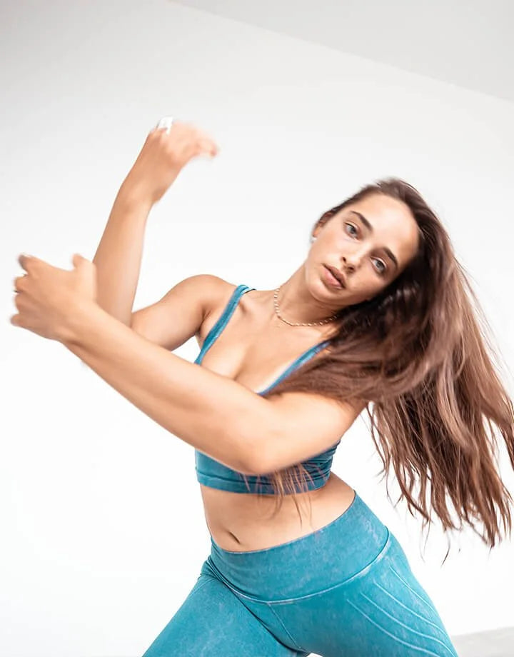A woman in a teal blue sports bra and matching high-waisted leggings poses dynamically against a clean white background.