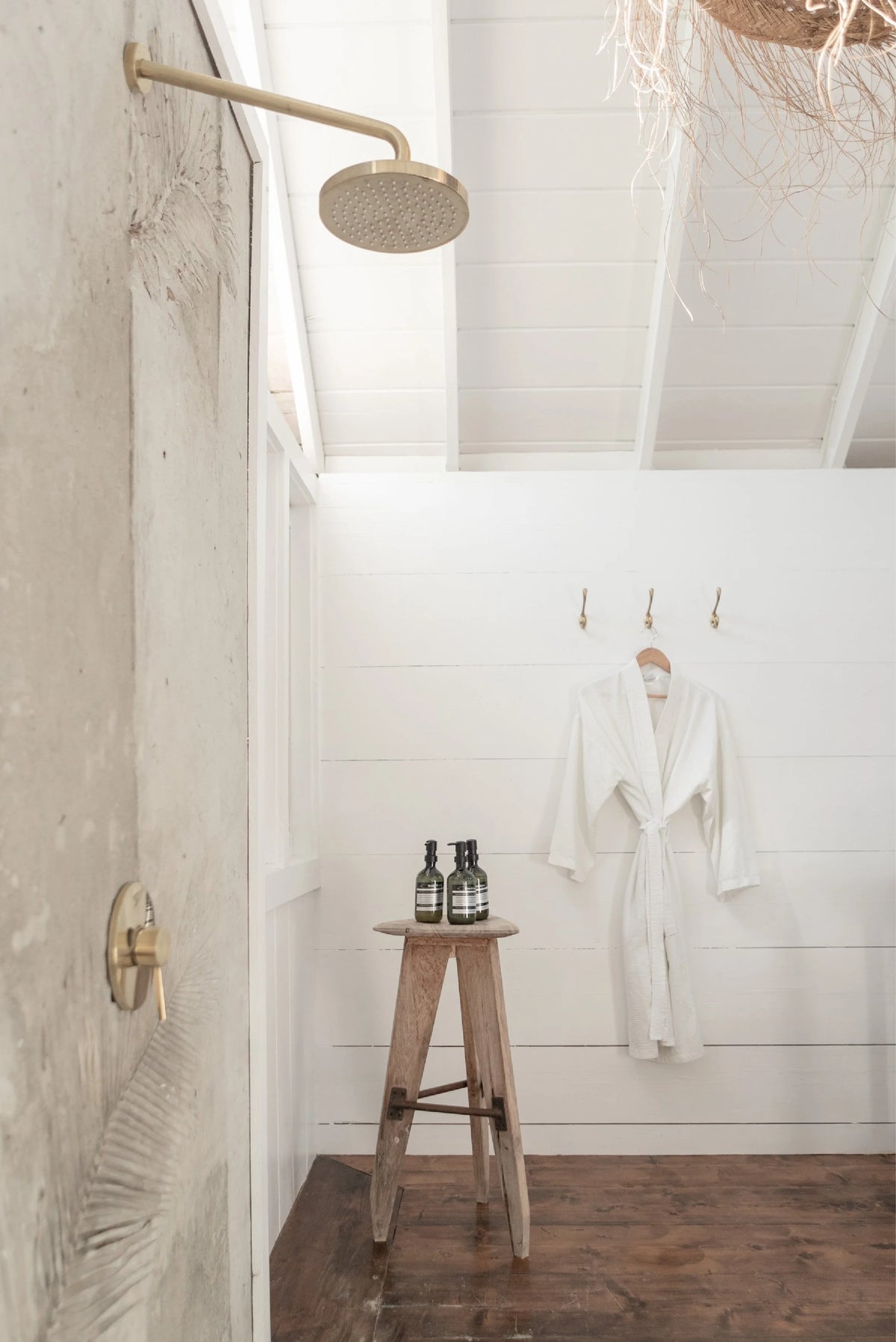 A rustic modern bathroom interior with a white shiplap wall, a white robe hanging on hooks, a small wooden stool with two dark bottles, and a brass shower head mounted on a textured concrete wall.
