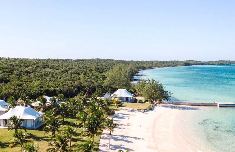Aerial view of a tropical beachfront resort with white tents, palm trees, a sandy shore, and turquoise ocean water under a clear blue sky.