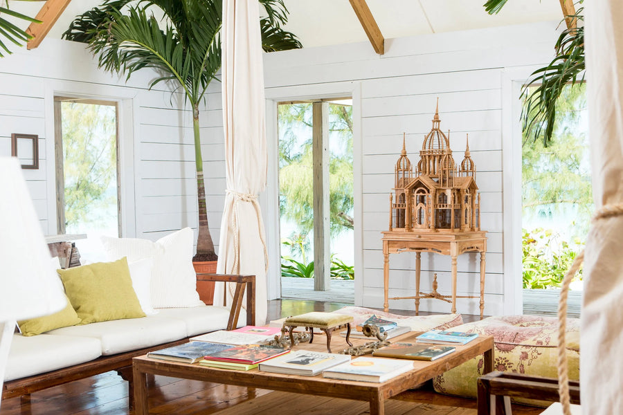 A bright, tropical-style living room with white shiplap walls, large windows, and wooden furniture. A white sofa with yellow-green pillows sits on the left, a wooden coffee table with magazines is in the foreground, and a decorative wooden church tower model stands on a stand against the back wall.