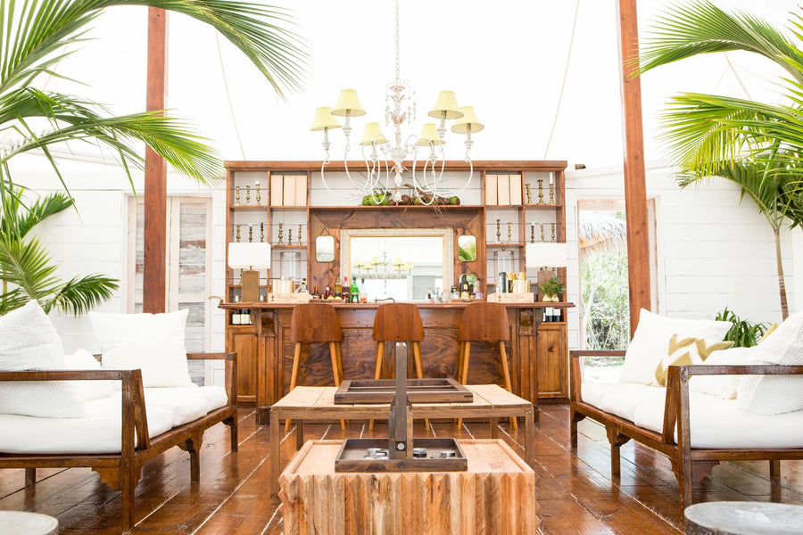 A tropical-style lounge bar interior with wooden furniture, white cushions, palm leaves, and a large chandelier in a bright, airy room.