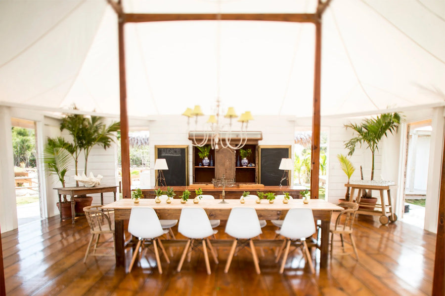 A rustic wooden dining table with six white chairs is set inside a large white canvas event tent with hardwood floors and potted plants.