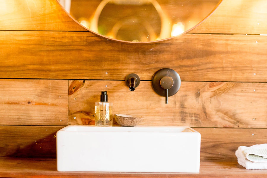 A white rectangular vessel sink with a matte black wall-mounted faucet and controls, set against a rustic wood plank wall. A small clear bottle and a folded white towel sit nearby.