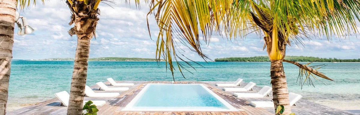 A tropical beach scene with palm trees framing a small pool, white lounge chairs, turquoise water, and a distant shoreline under a partly cloudy sky.