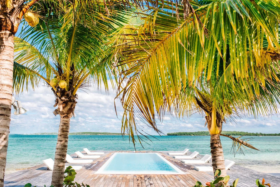A tropical beach scene with palm trees framing a small pool, white lounge chairs, turquoise water, and a distant shoreline under a partly cloudy sky.