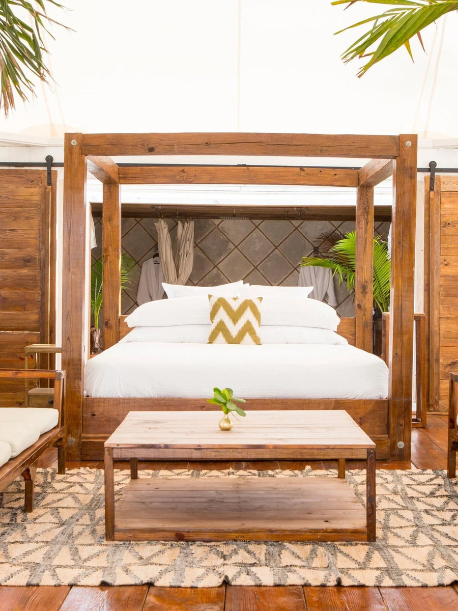 A rustic wooden four-poster bed with white bedding and a gold chevron accent pillow, styled in a tropical lodge setting with a matching wooden coffee table and patterned rug.
