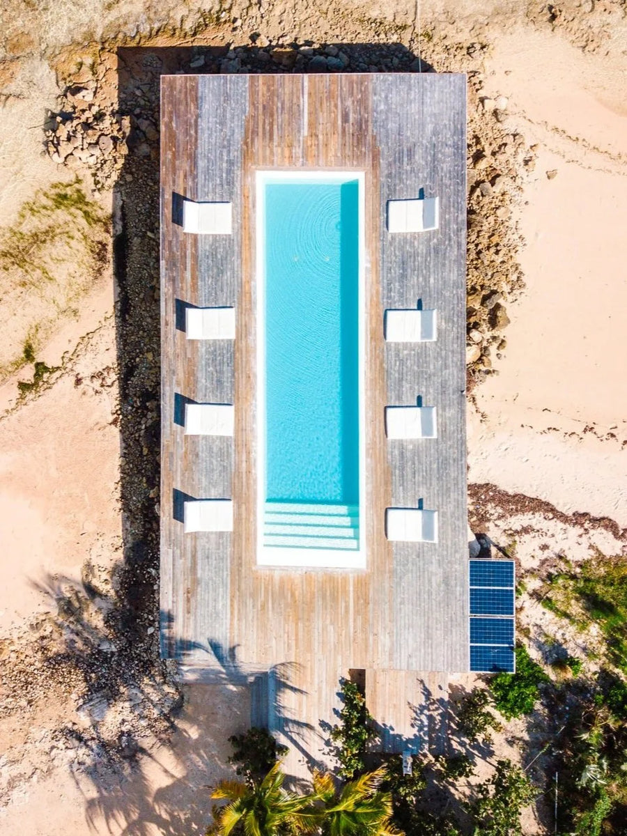 Aerial view of a modern rectangular villa with a long turquoise swimming pool, wooden deck, white lounge chairs, sandy beach, and rocky shoreline.