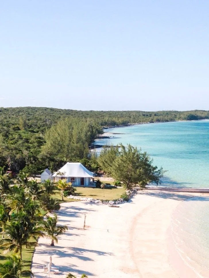 A scenic tropical beach with pale sand, turquoise water, a line of green trees, and a white tent-like structure near the shoreline under a clear blue sky.