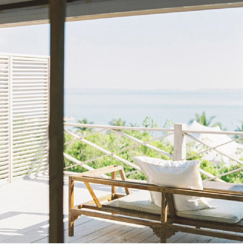 A wooden outdoor lounge chair with a white cushion and pillow sits on a sunlit deck overlooking a bright ocean view with greenery and umbrellas in the background.