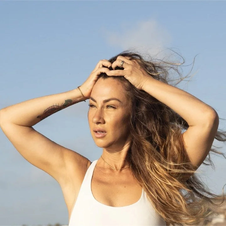 A woman with long, wind-blown brown hair wearing a white tank top, posing outdoors against a clear blue sky with her hands in her hair.