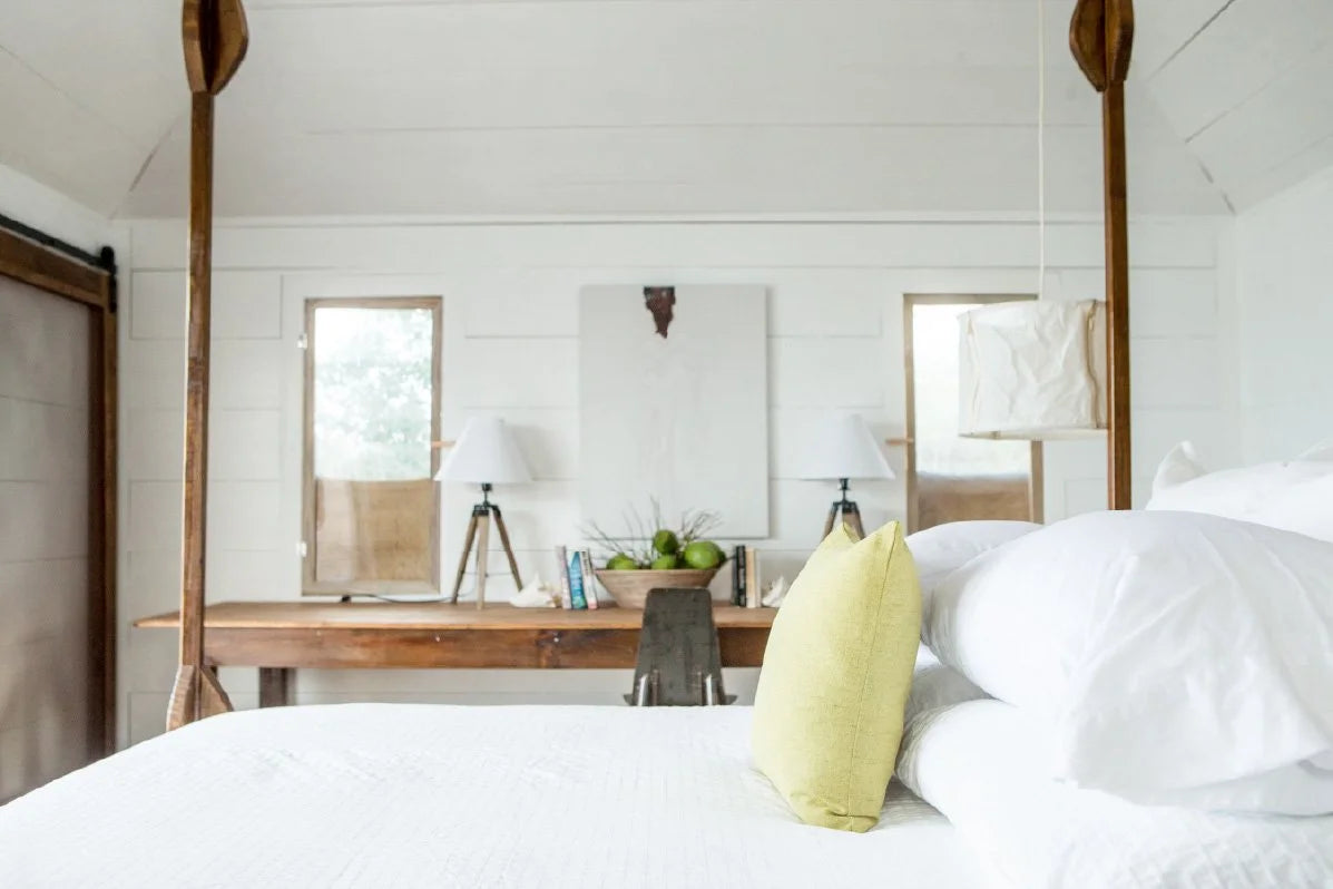 A bright, modern bedroom featuring a white canopy bed in the foreground with a yellow accent pillow, set against a white shiplap wall and a rustic wooden console table.