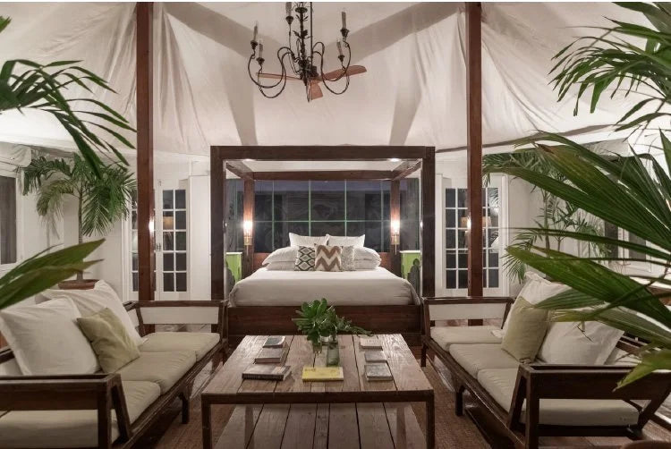 A luxury tropical resort bedroom and lounge area inside a large white canvas pavilion. The bedroom features a dark wood four-poster bed with white linens, flanked by nightstands and lamps.