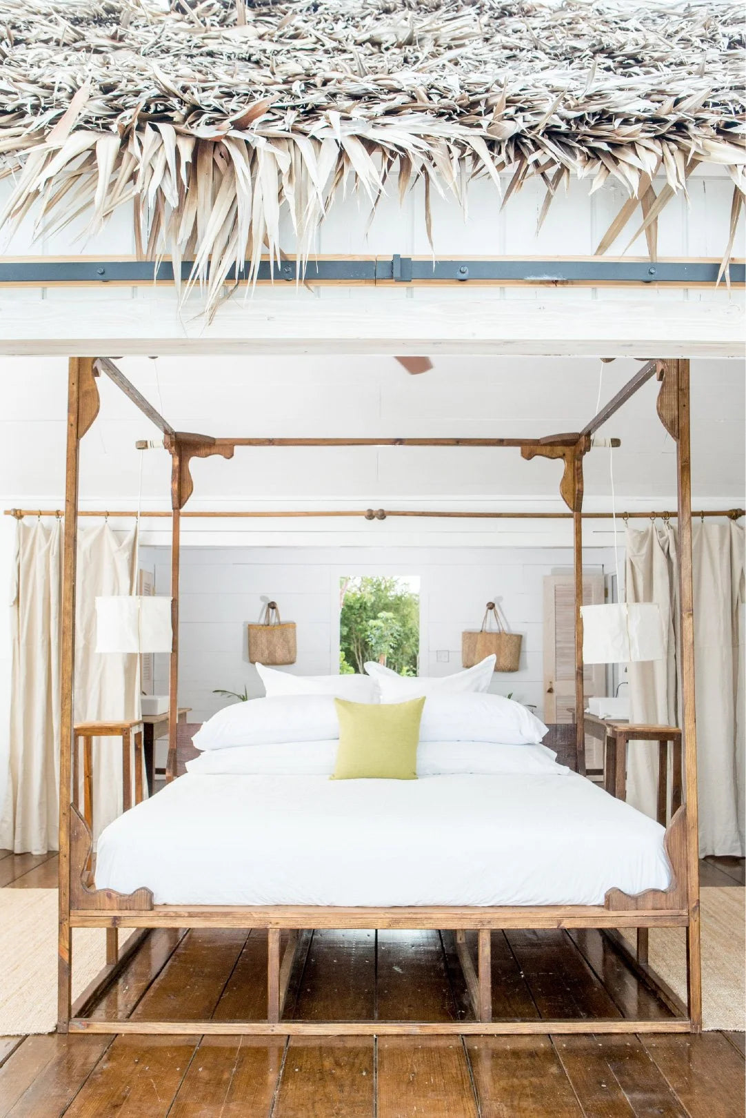 A rustic wooden four-poster bed with white bedding and a single light green accent pillow, set under a thatched roof in a bright, airy bedroom.