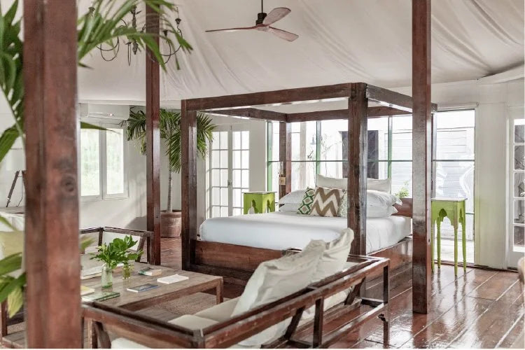 A rustic bedroom interior with a large wooden four-poster bed, matching wooden bench and table, white bedding, green accent chairs, and a white canvas tent ceiling with exposed wooden posts.