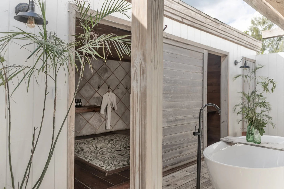 Outdoor bathroom scene with a freestanding white bathtub, dark faucet, wooden sliding door, tiled shower area, and tall palm plants against a white wall.