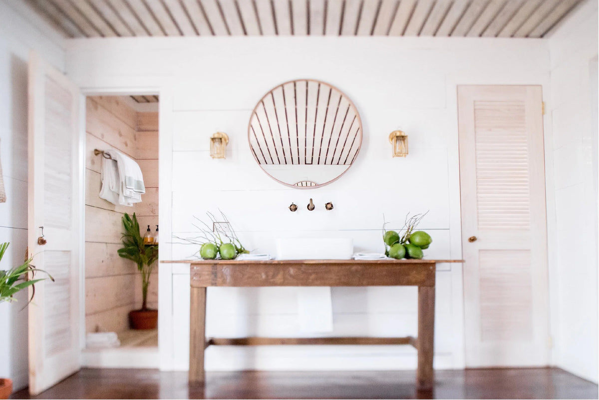 A rustic wooden console table in a bright, modern bathroom with white shiplap walls, a round wooden fan mirror, and green coconuts on top.
