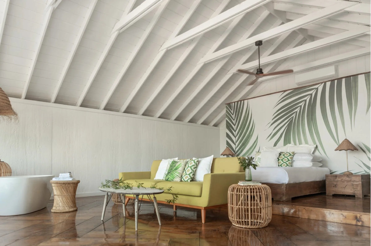 A bright, modern bedroom and living area with a green sofa, tropical leaf wall mural, wooden ceiling beams, and a large white tub on a wooden floor.