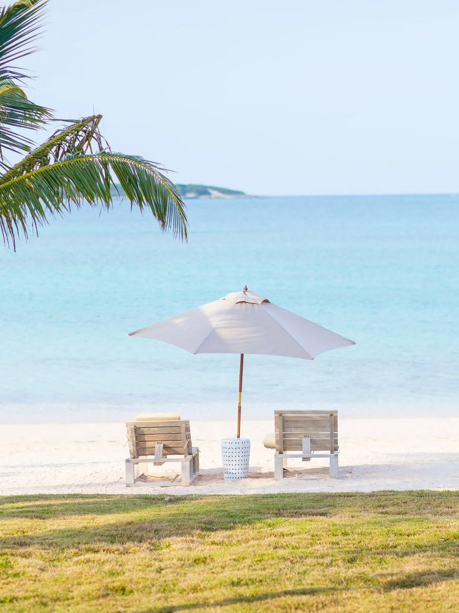 A white beach umbrella with two wooden lounge chairs on a white sand beach, with turquoise ocean water and a palm tree in the background.