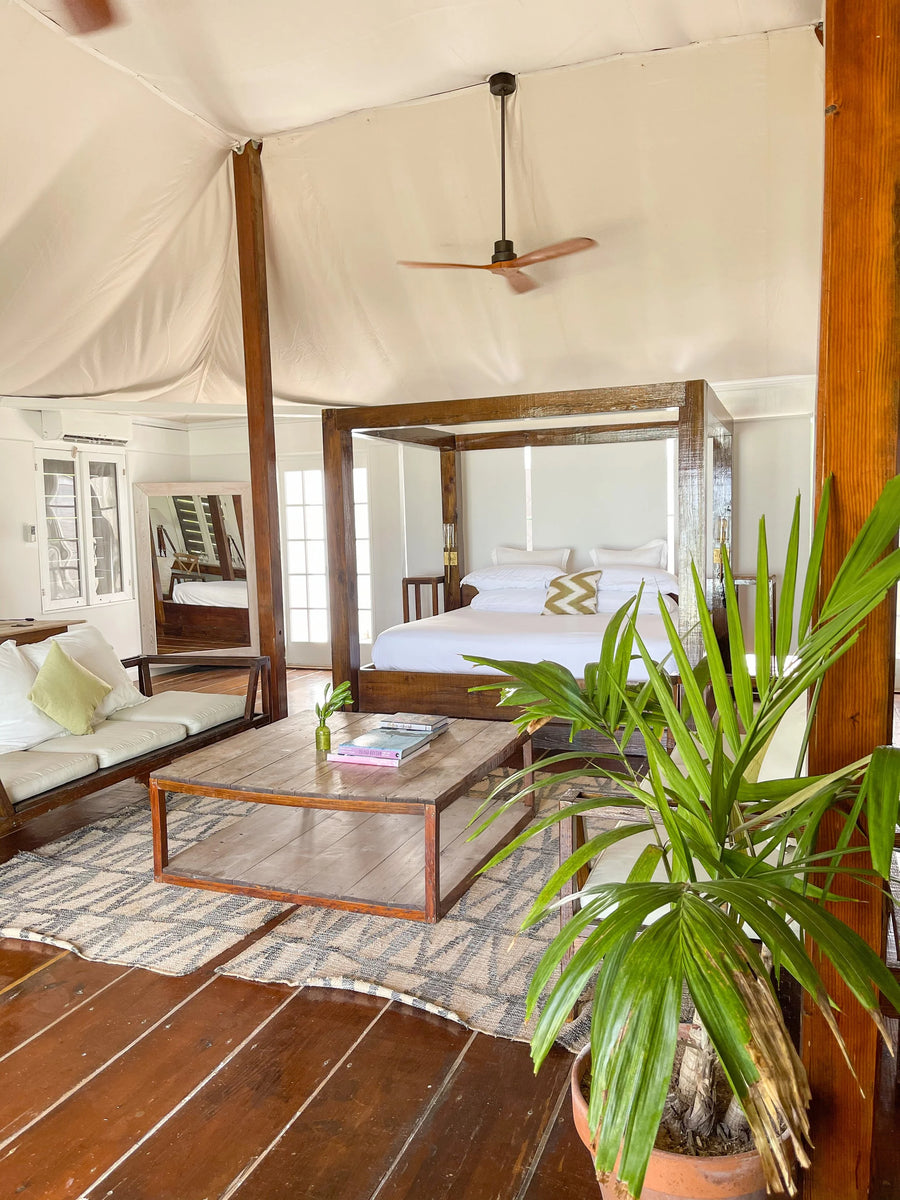 A rustic luxury tent-style bedroom with a wooden four-poster bed, white bedding, a low wooden coffee table, patterned rugs, a bench with pillows, and a large potted green plant in the foreground.