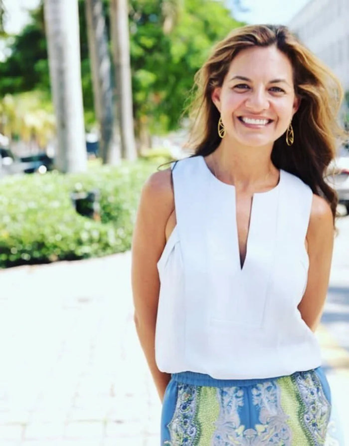 A smiling woman standing outdoors on a sunny sidewalk, wearing a sleeveless white top with a split neckline and a blue, green, and white patterned skirt.