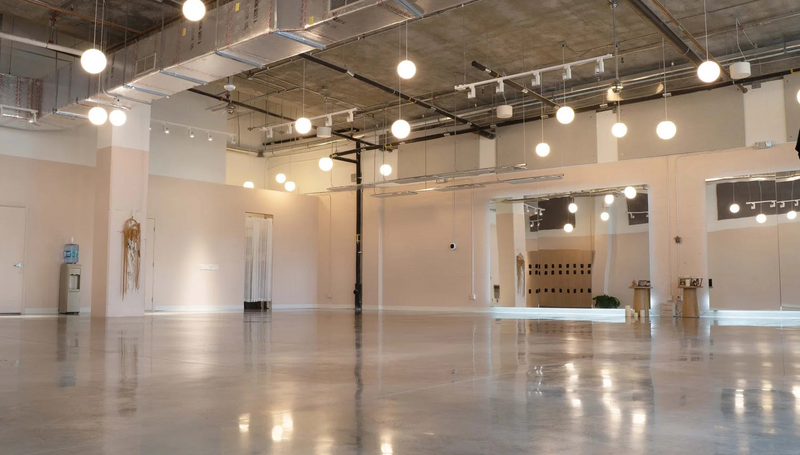 An empty industrial loft space with a polished concrete floor, high ceiling with exposed ducts and hanging spherical pendant lights, and white walls with display openings.