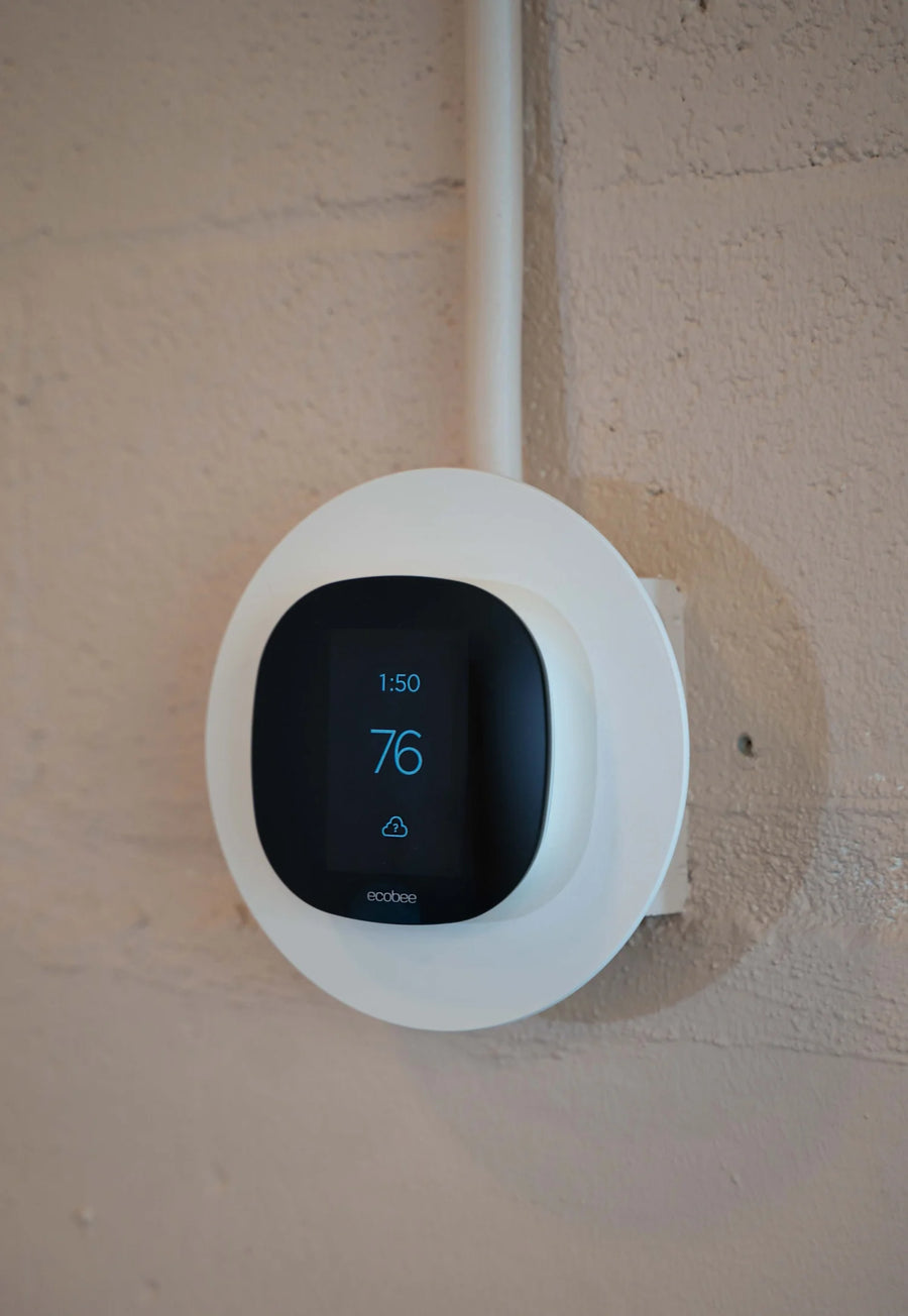 A white Ecoblast smart thermostat mounted on a textured beige wall, showing a black display screen with blue digital temperature and time indicators.