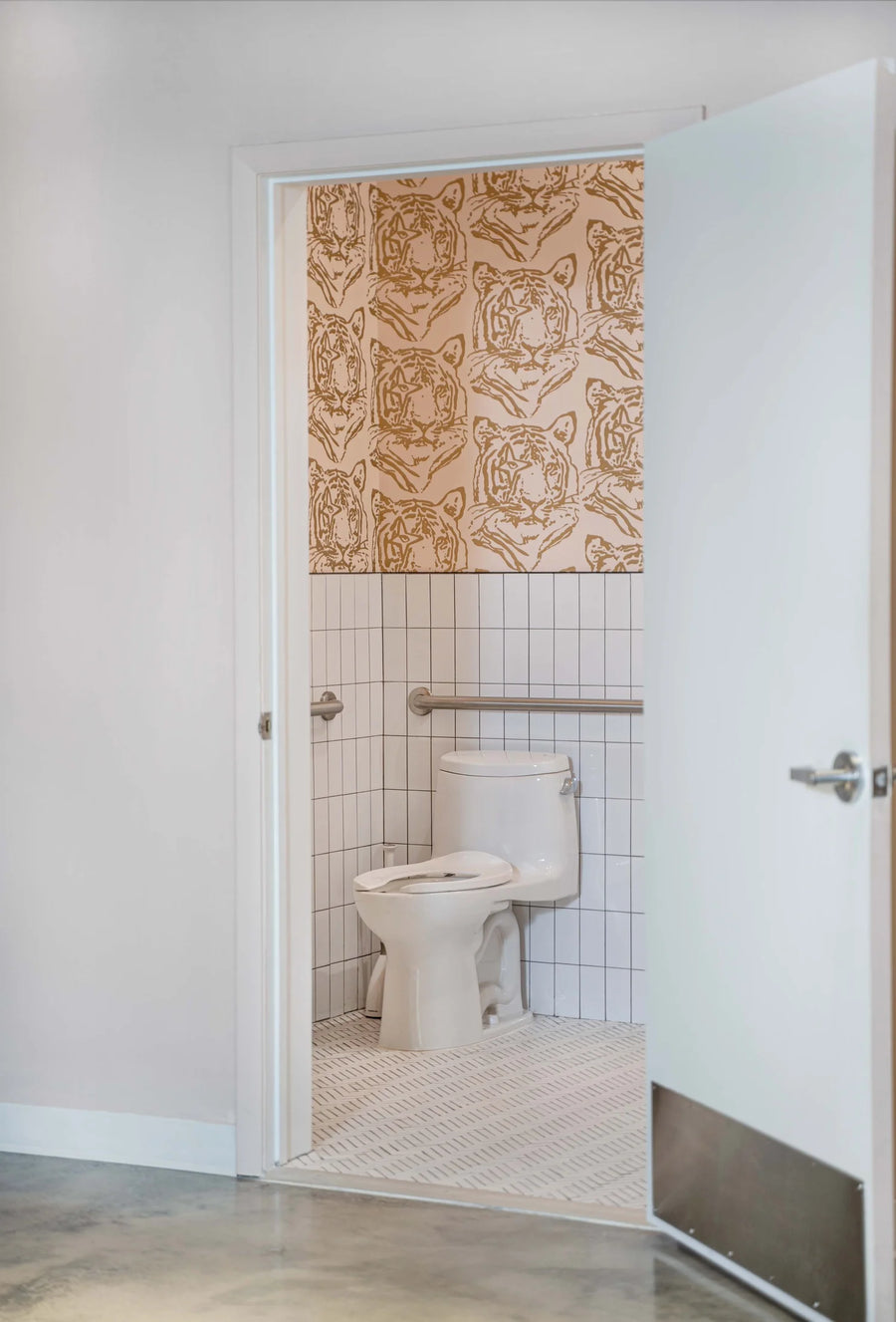 A bathroom interior with a white toilet, white tiled walls, a patterned beige and brown tiger-print curtain hanging above the toilet, and an open white door on the right.