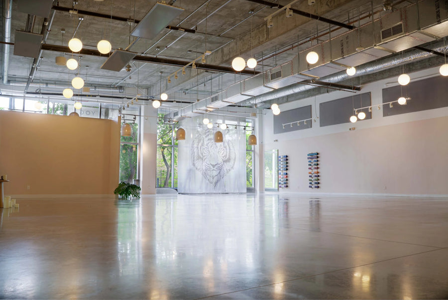 An empty, spacious industrial loft or art gallery with a polished concrete floor, high ceiling with exposed ducts and multiple hanging globe lights, and a large white wall in the distance featuring a faint animal illustration.