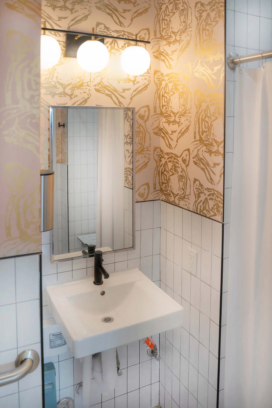 A modern bathroom vanity featuring a white wall-mounted sink, a black faucet, a rectangular mirror, and a light fixture with three glowing globe bulbs. The walls are decorated with white square tiles and a distinctive beige and gold patterned wallpaper.