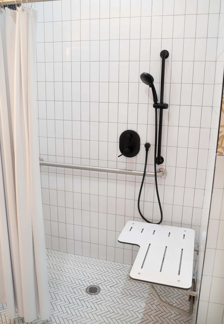 A modern walk-in shower area with white square tiles, a black handheld showerhead and controls, a white fold-down seat, and a white shower curtain on the left.