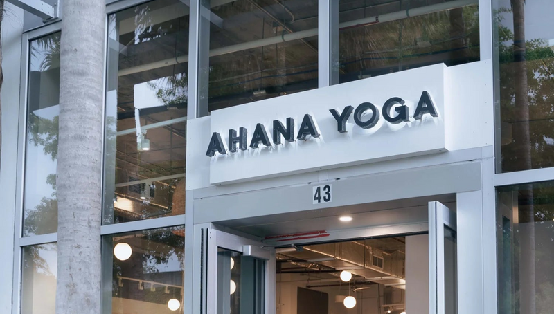 The entrance to Ahana Yoga studio, featuring glass double doors with silver frames, a white sign above the door with black lettering, and a modern glass facade. A tree trunk is visible on the left foreground.