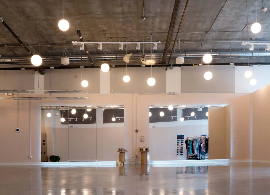 A large, empty modern retail space or studio with a polished concrete floor, high ceiling with exposed ductwork, and multiple spherical pendant lights hanging at varying heights. Two glass-walled back-of-house or fitting rooms are visible in the background, along with clothing racks inside.