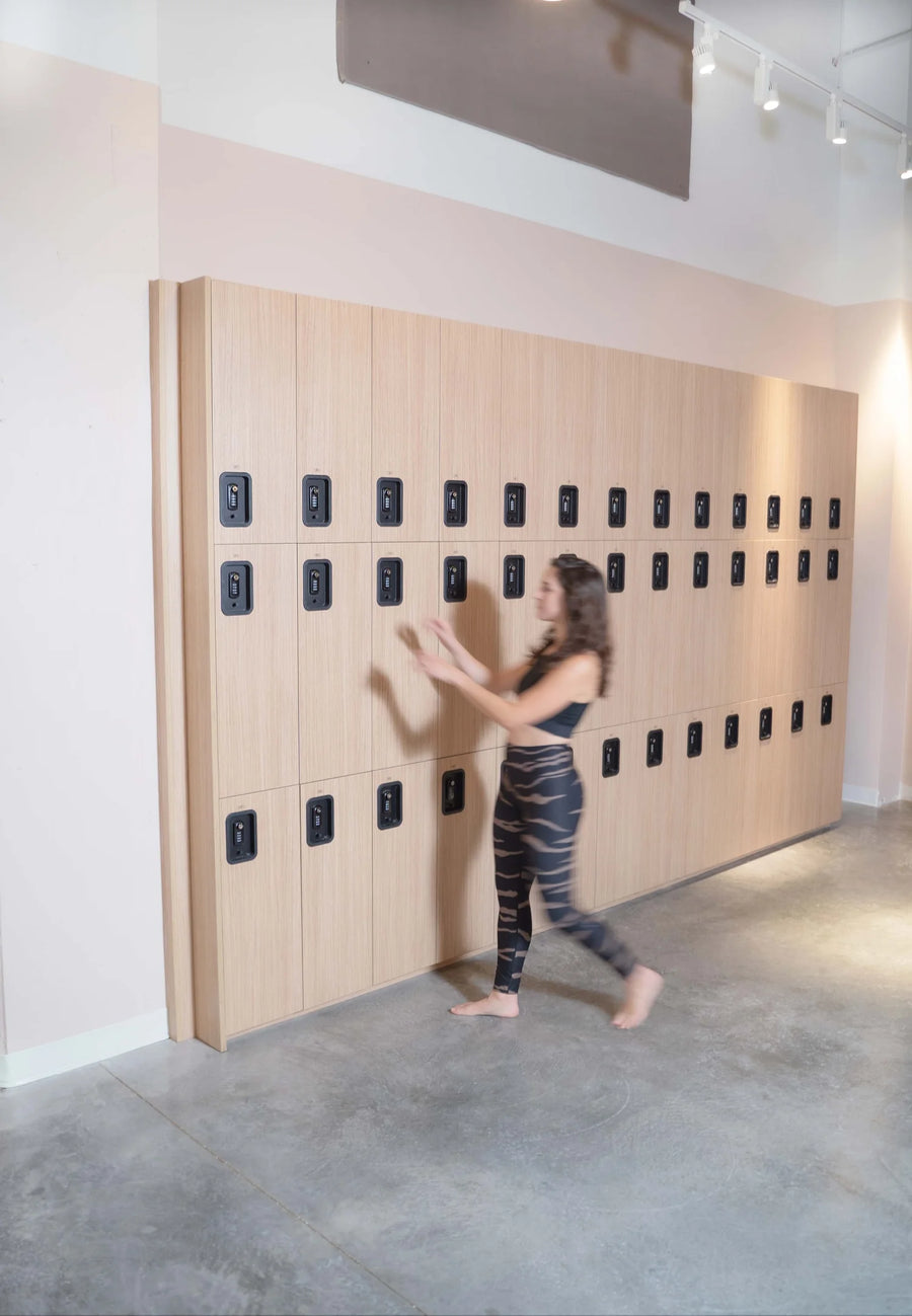 A woman in a black sports bra and patterned leggings walks past a large wall-mounted wooden locker system with black electronic lock panels in a modern indoor space with concrete floors.