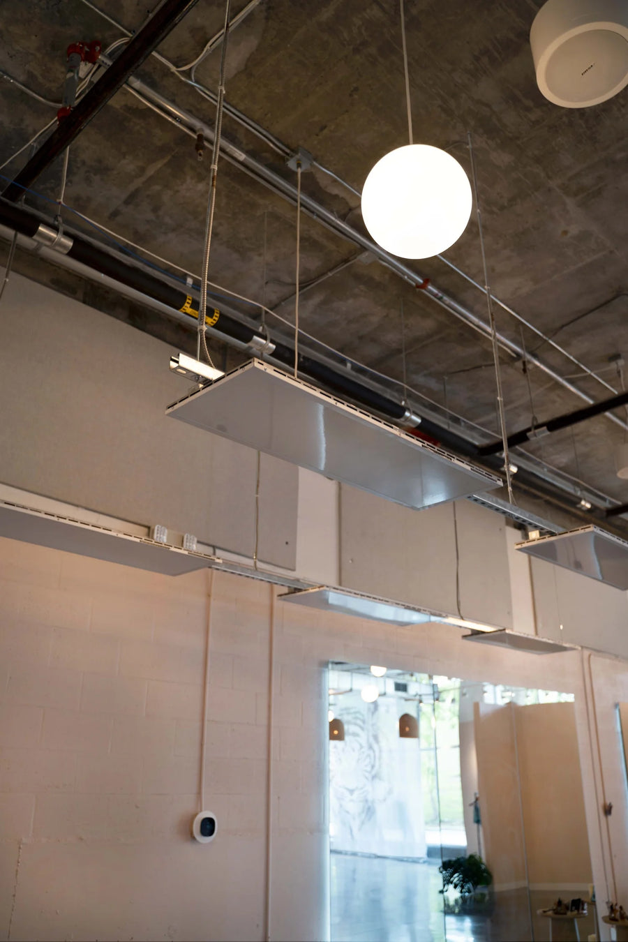 Industrial ceiling-mounted ventilation ducts and metal grilles in a modern indoor space with concrete ceiling, hanging spherical pendant lights, and light-colored walls.