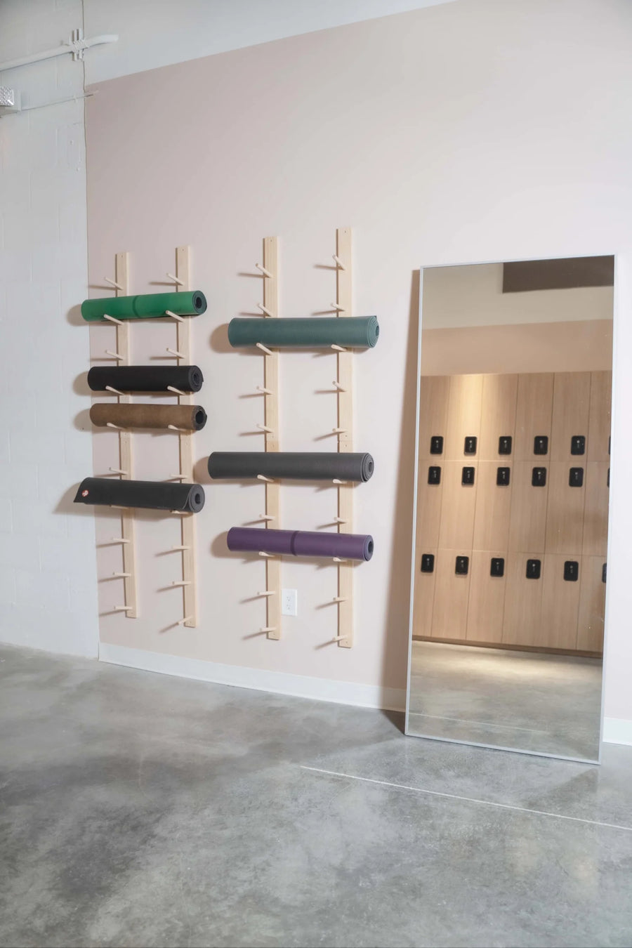 A minimalist interior space with a large floor mirror and wall-mounted wooden racks holding several rolled exercise or yoga mats in green, black, tan, gray, and purple. The room has a pale blush wall, gray concrete floor, and light wood cabinetry reflected in the mirror.