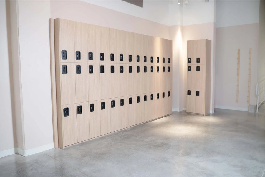A set of modern light wood locker cabinets with black electronic lock panels, installed on a wall in a minimalist room with concrete floors.