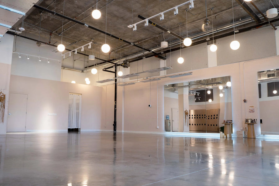 An empty modern art gallery or studio with a polished concrete floor, high ceiling with exposed ductwork and track lighting, and multiple hanging spherical pendant lights.