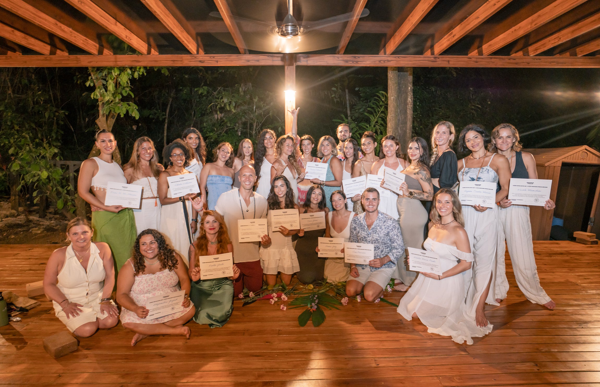 A large group of women and a few men posing together on a wooden deck under a covered pavilion at night, each holding a certificate. The background shows trees and warm ambient lighting.