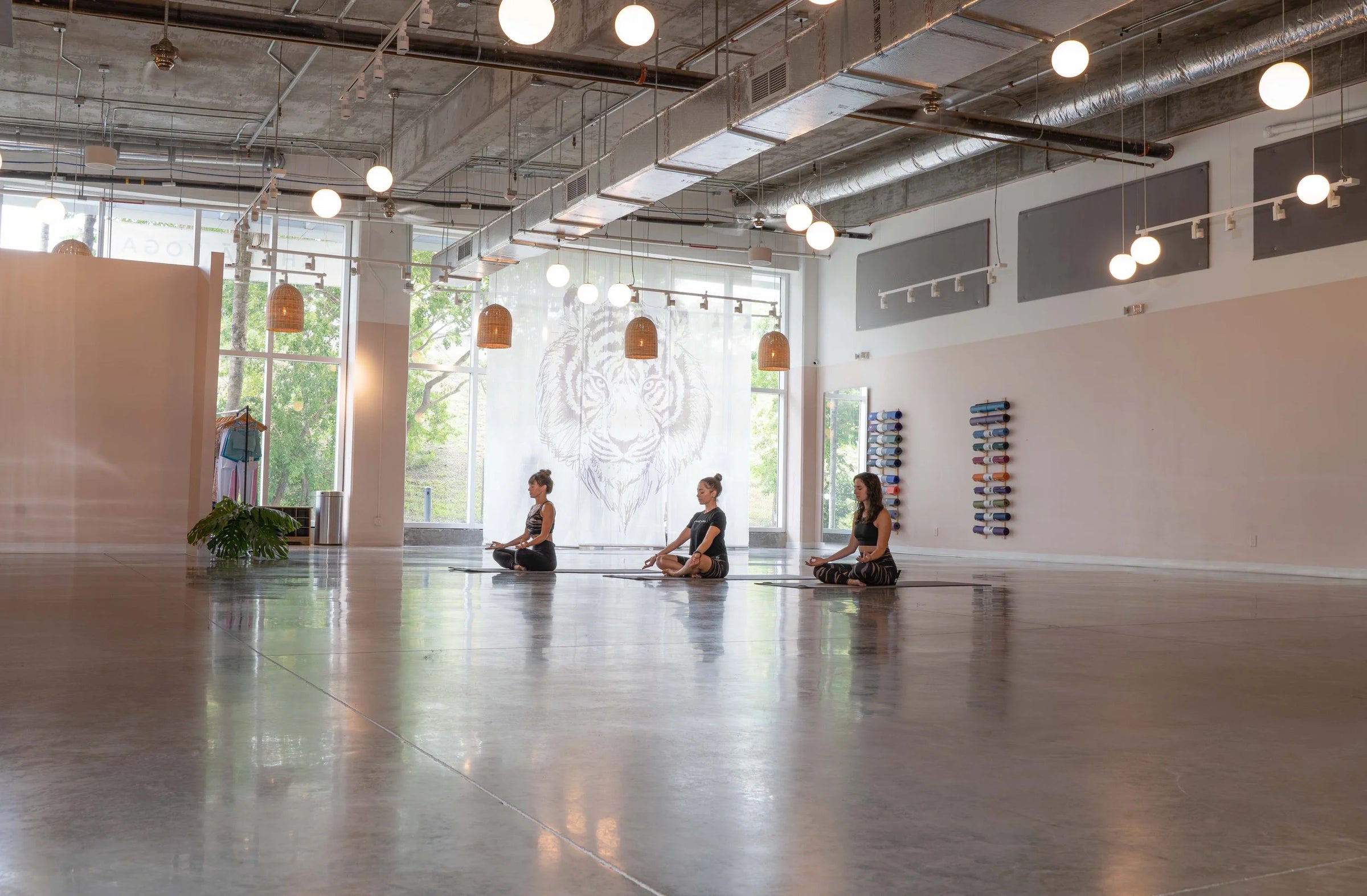 Three people sitting cross-legged on yoga mats in a spacious, modern studio with high ceilings, industrial ductwork, and warm pendant lights. A large tiger illustration is visible on the back wall.
