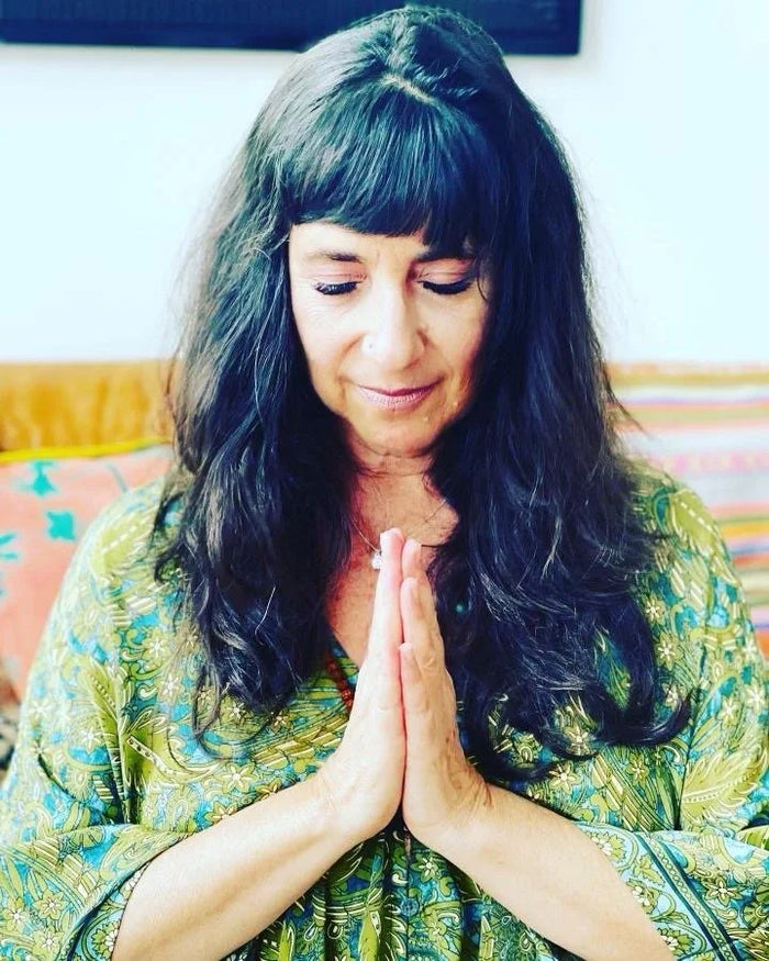 A woman with long dark hair and bangs, wearing a green and yellow patterned top, with her hands pressed together in a prayer pose.