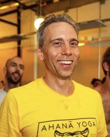 A smiling man wearing a bright yellow t-shirt with 'AHANA YOGA' printed on the front, standing in what appears to be a yoga studio or gym.